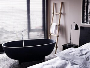 A modern bathroom with a freestanding bathtub, a wooden ladder, a bedside table with a lamp, and a bed with white linens.