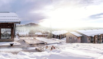 Un centro turístico nevado con cabañas de madera, una bañera de hidromasaje y vistas a una cadena montañosa boscosa.