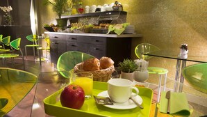A breakfast setup with a tray containing an apple, orange juice, and pastries, alongside a cup and saucer, in a modern dining area with glass chairs and a buffet in the background. - Le Fabe Hotel (Paris)