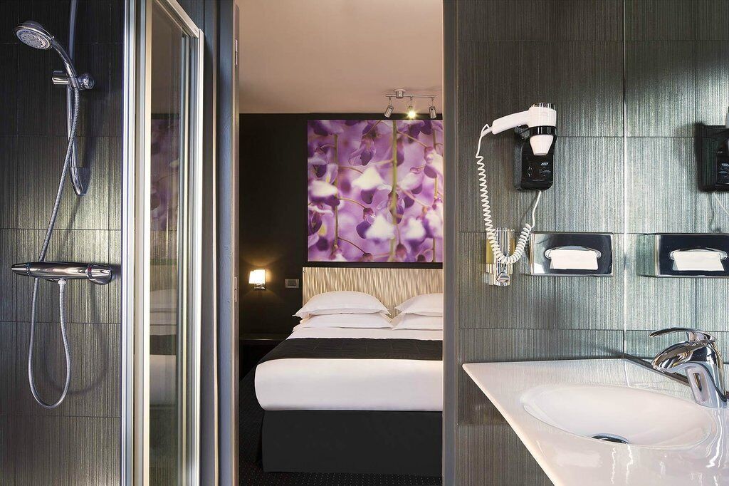 Een moderne hotelbadkamer met een glazen douchecabine, een groot bed met witte lakens, een muurbevestigde haardroger en een wastafel met spiegel.