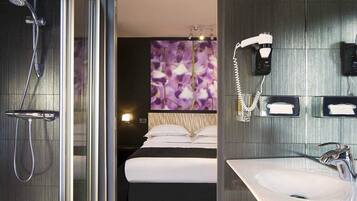 Een moderne hotelbadkamer met een glazen douchecabine, een groot bed met witte lakens, een muurbevestigde haardroger en een wastafel met spiegel.