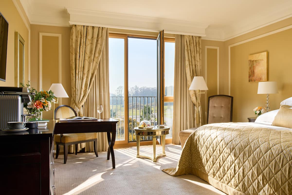 A hotel room with a large bed, a desk with a chair, a small table with a lamp, and a view of the outdoors through a large window.