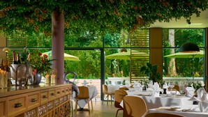 Un restaurante con mesas tanto en el interior como en el exterior, amplias ventanas y plantas.