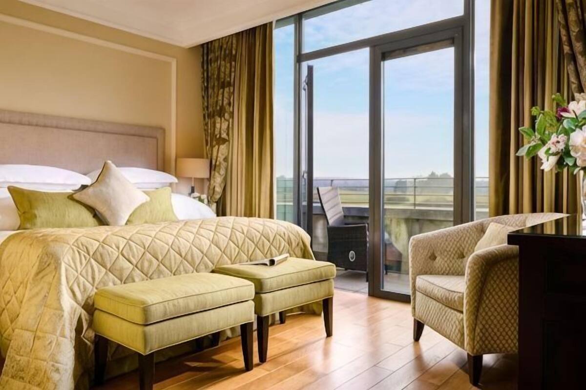 A hotel room with a bed, two ottomans, a chair, a desk, and a view through a large window.