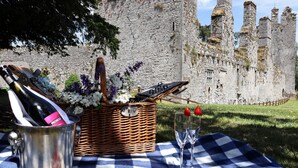 Un montaje para picnic con una cesta, una botella de vino y copas sobre un mantel a cuadros, frente a una pared de piedra histórica.