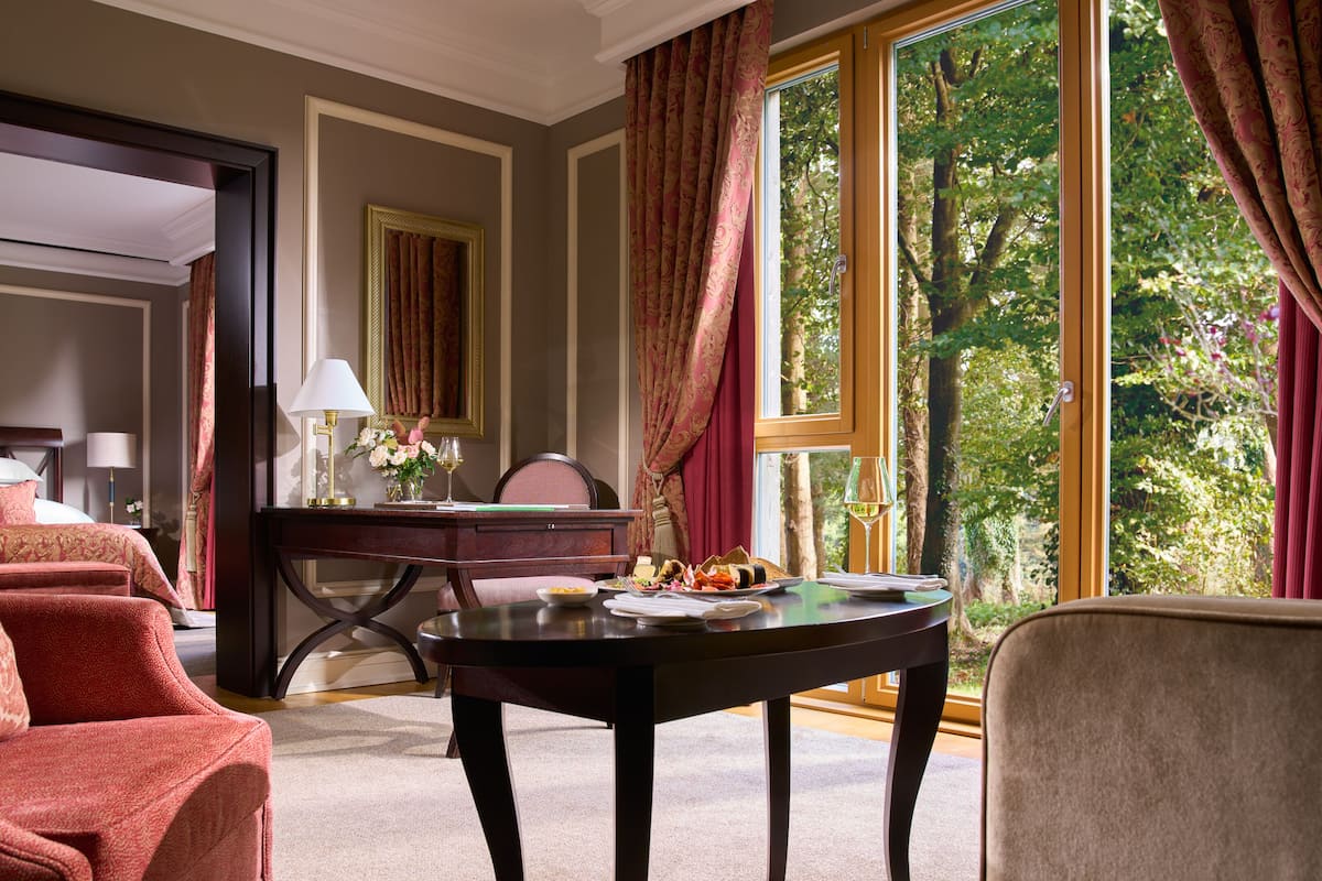 A hotel room with a bed, a desk with a lamp, a dining table with food, and large windows with curtains.