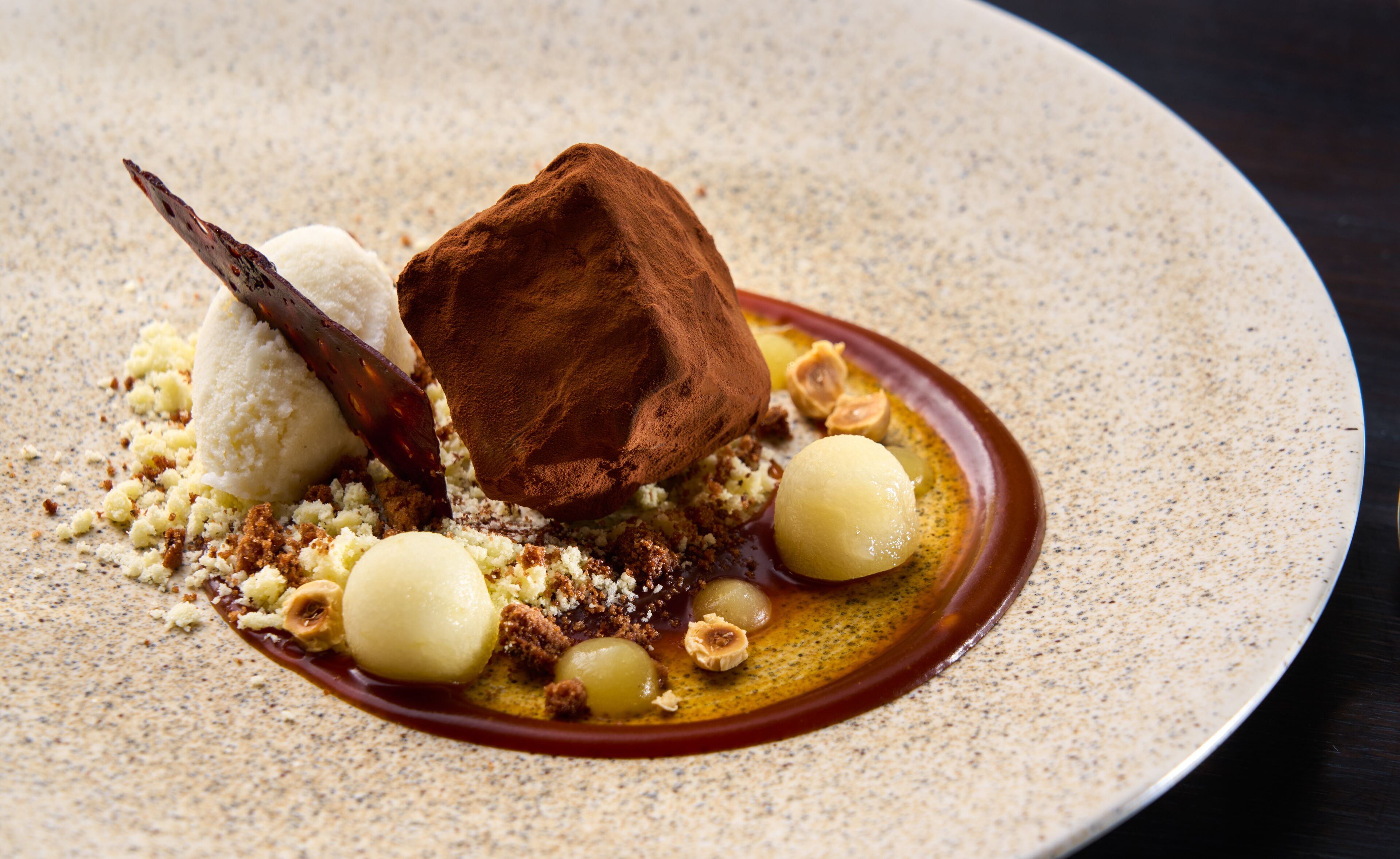 Sobremesa com pedaço de chocolate, sorvete e nozes sobre um prato texturizado.
