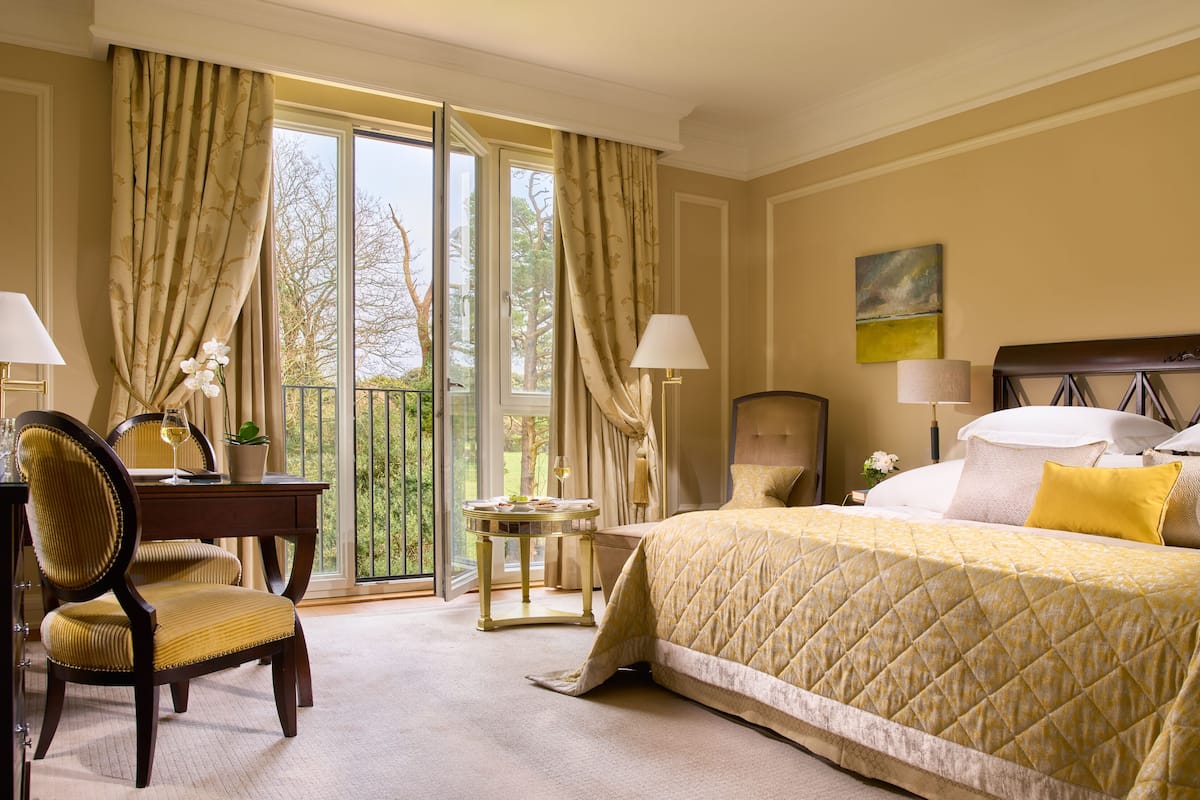A hotel room with a large bed, two bedside tables, a desk with a chair, and a view of the outdoors through large windows.