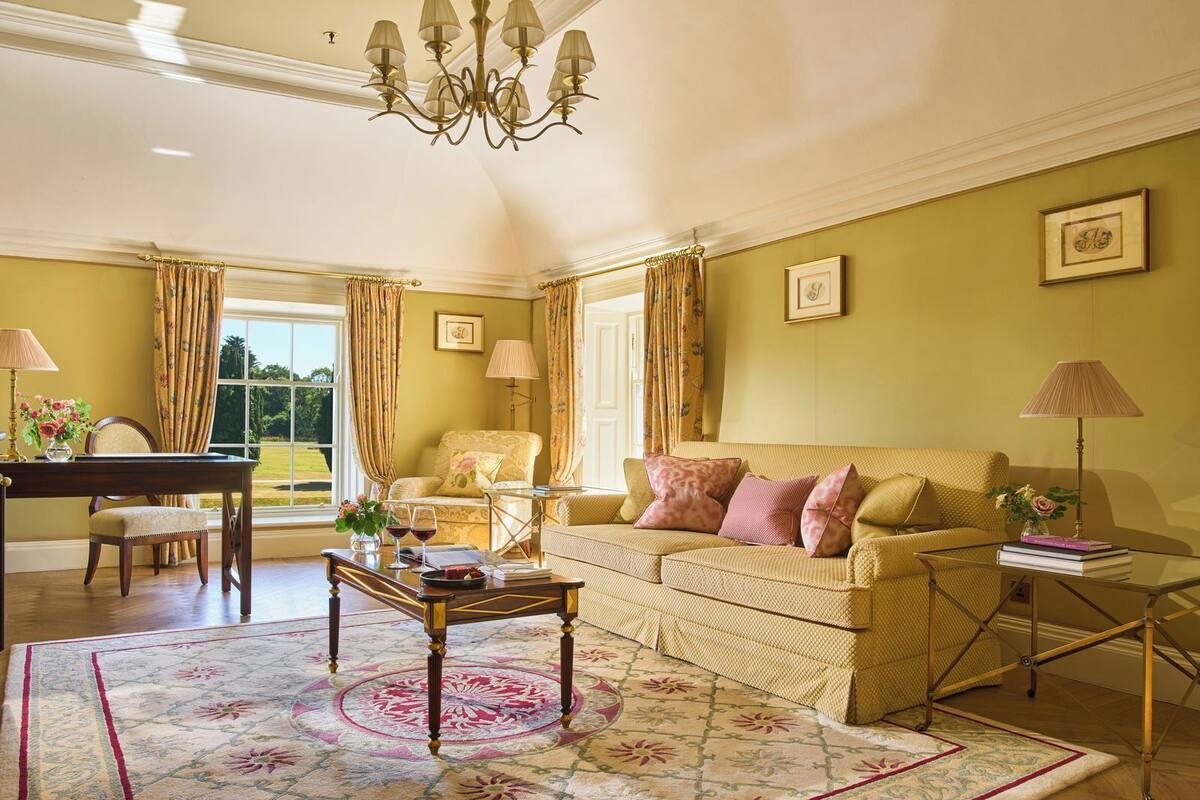 A well-lit living room with a sofa, armchair, and a dining table with chairs. There is a chandelier hanging from the ceiling and a large window with curtains.