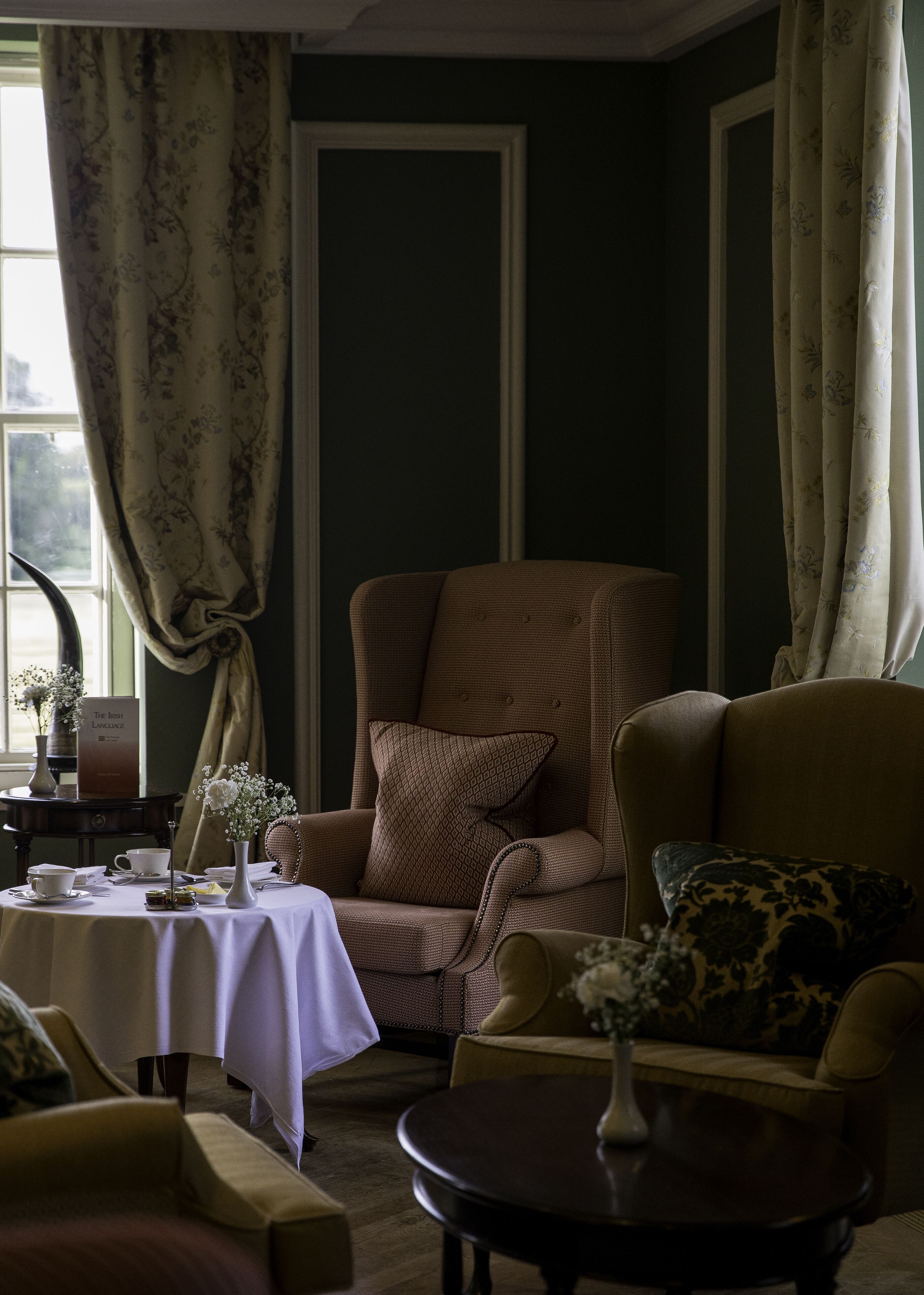 Una habitación con una pared verde oscuro, un sillón marrón con un cojín estampado, un sillón amarillo, una mesita redonda con mantel blanco y un florero con flores.