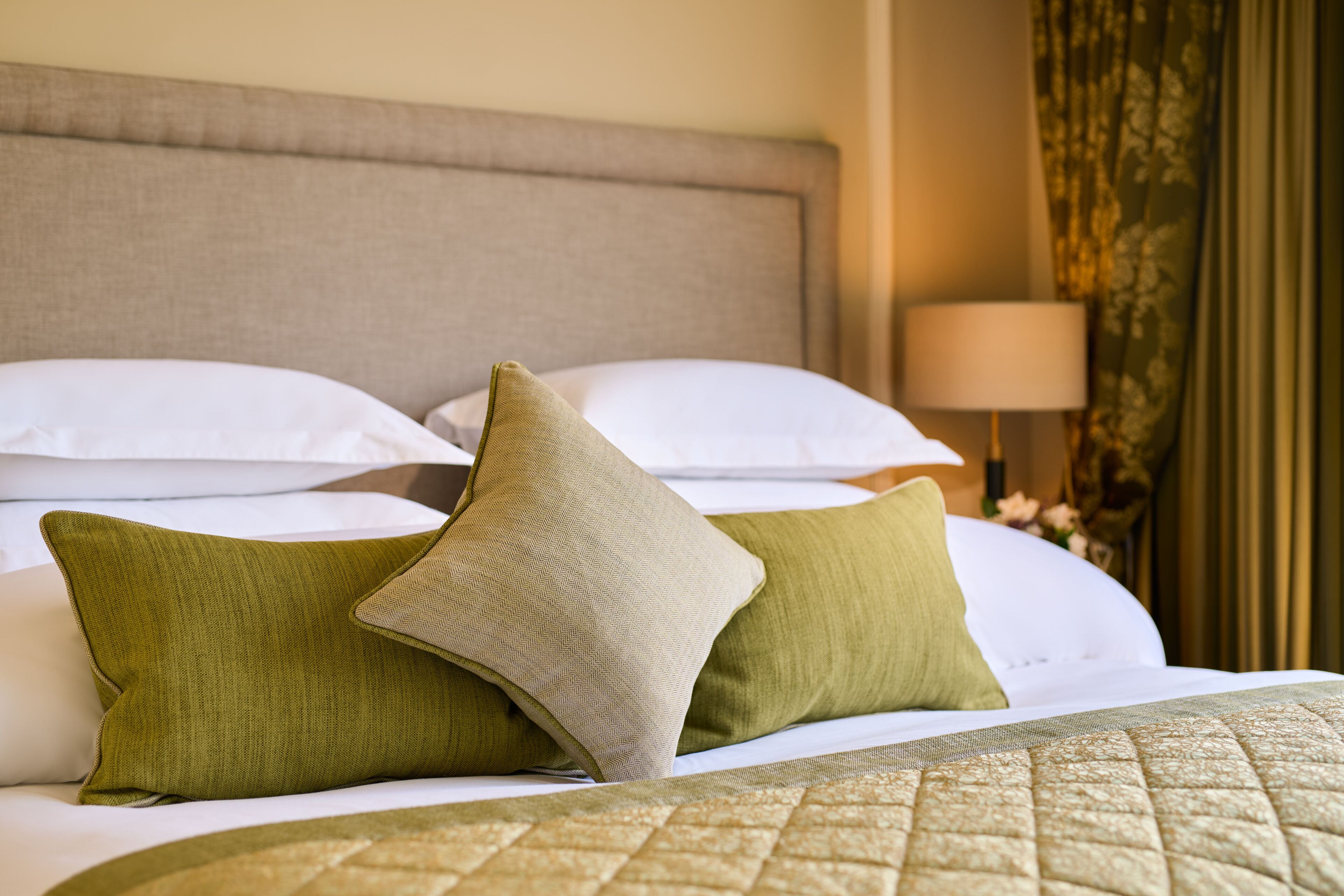 A neatly made bed with green and beige pillows, a quilted bedspread, and a lamp in the background.