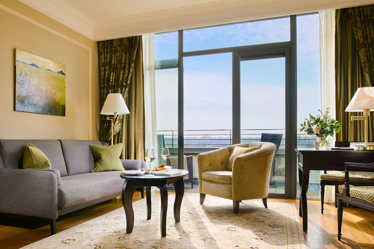 A modern hotel room with a grey sofa, a round table, a yellow armchair, and a large window offering a view of the city.