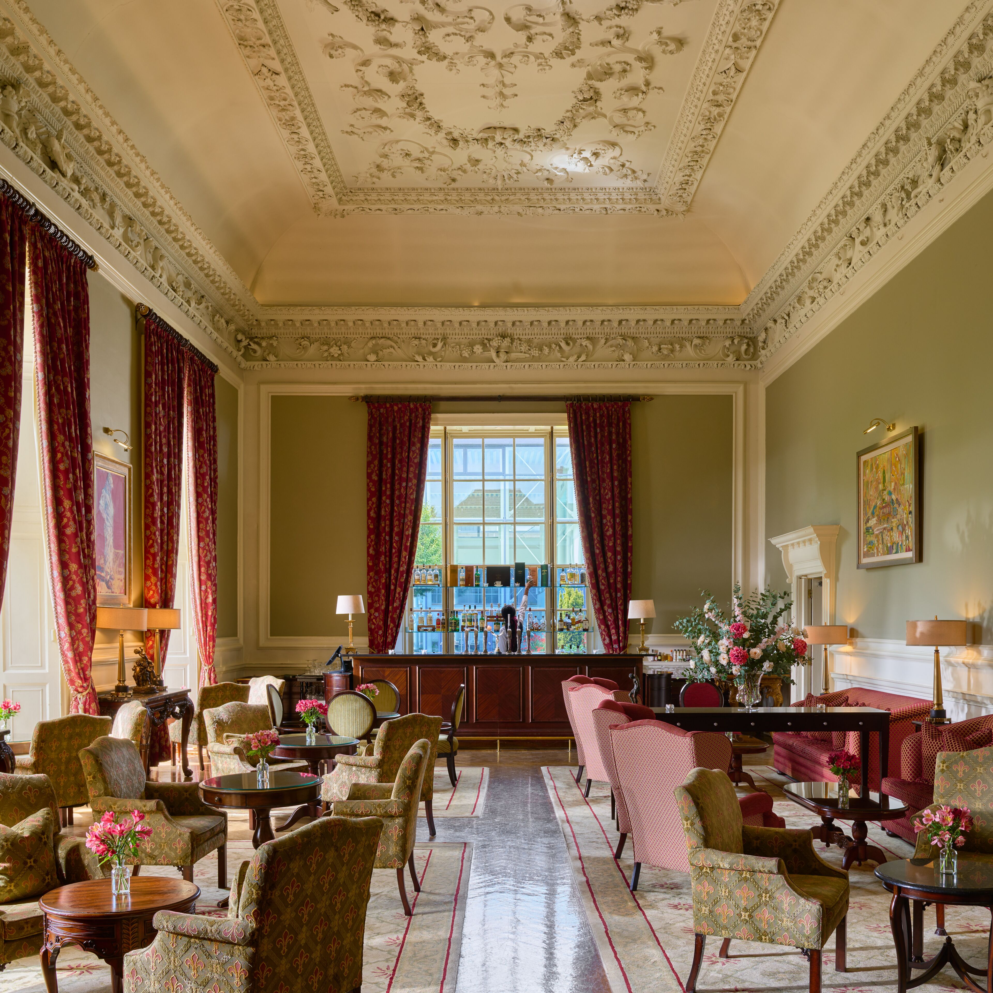 A grand room with ornate ceiling and floral wallpaper, featuring a bar area, multiple seating arrangements, and a large window with red curtains.