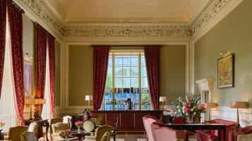 A grand room with ornate ceiling and floral wallpaper, featuring a bar area, multiple seating arrangements, and a large window with red curtains.