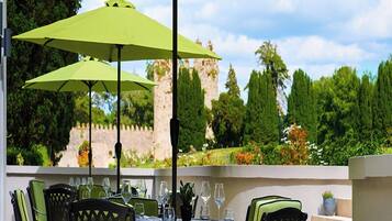 An outdoor dining area with tables and chairs, green umbrellas, and a view of a stone wall and trees.
