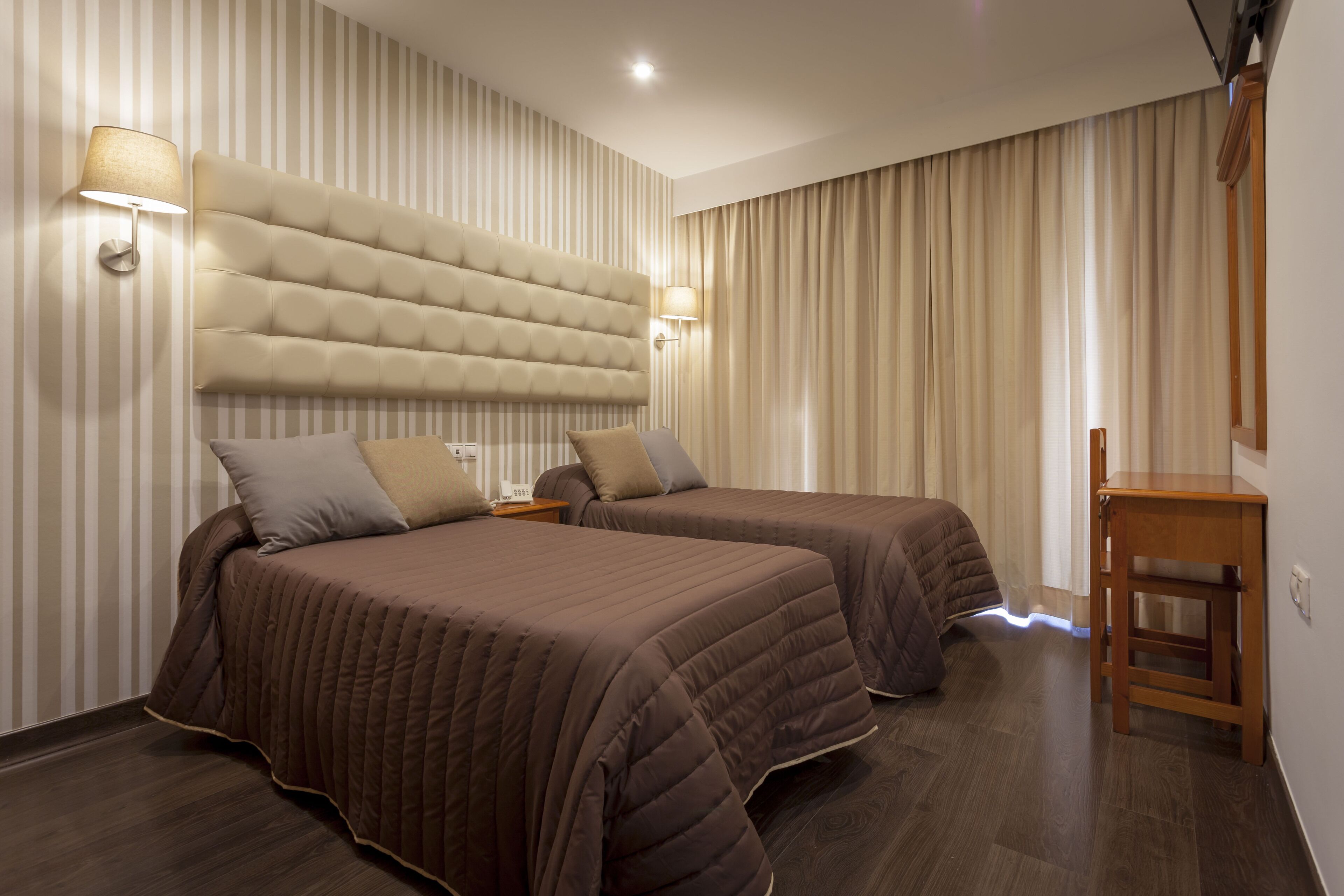 Ein Hotelzimmer mit zwei Betten, einem großen gepolsterten Kopfteil, Wandlampen und einem Holznachttisch.