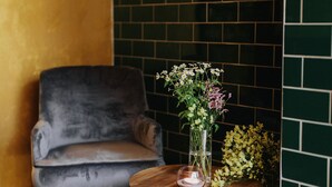 Una habitación con un sillón gris, una mesa redonda de madera con un florero, una alfombra estampada y una pared con azulejos verdes.