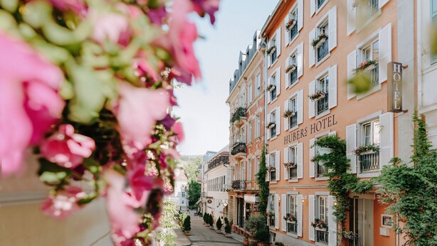 Un hotel con un letrero que dice "HUBER'S HOTEL" y un letrero de restaurante. El edificio tiene varios pisos con balcones y macetas de flores.