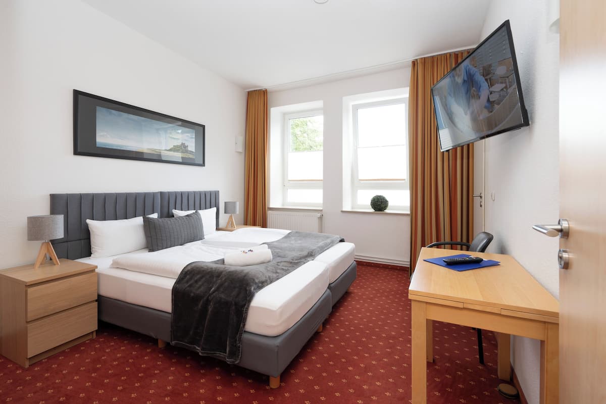 A hotel room with a bed, bedside tables, a desk, a television, and windows.