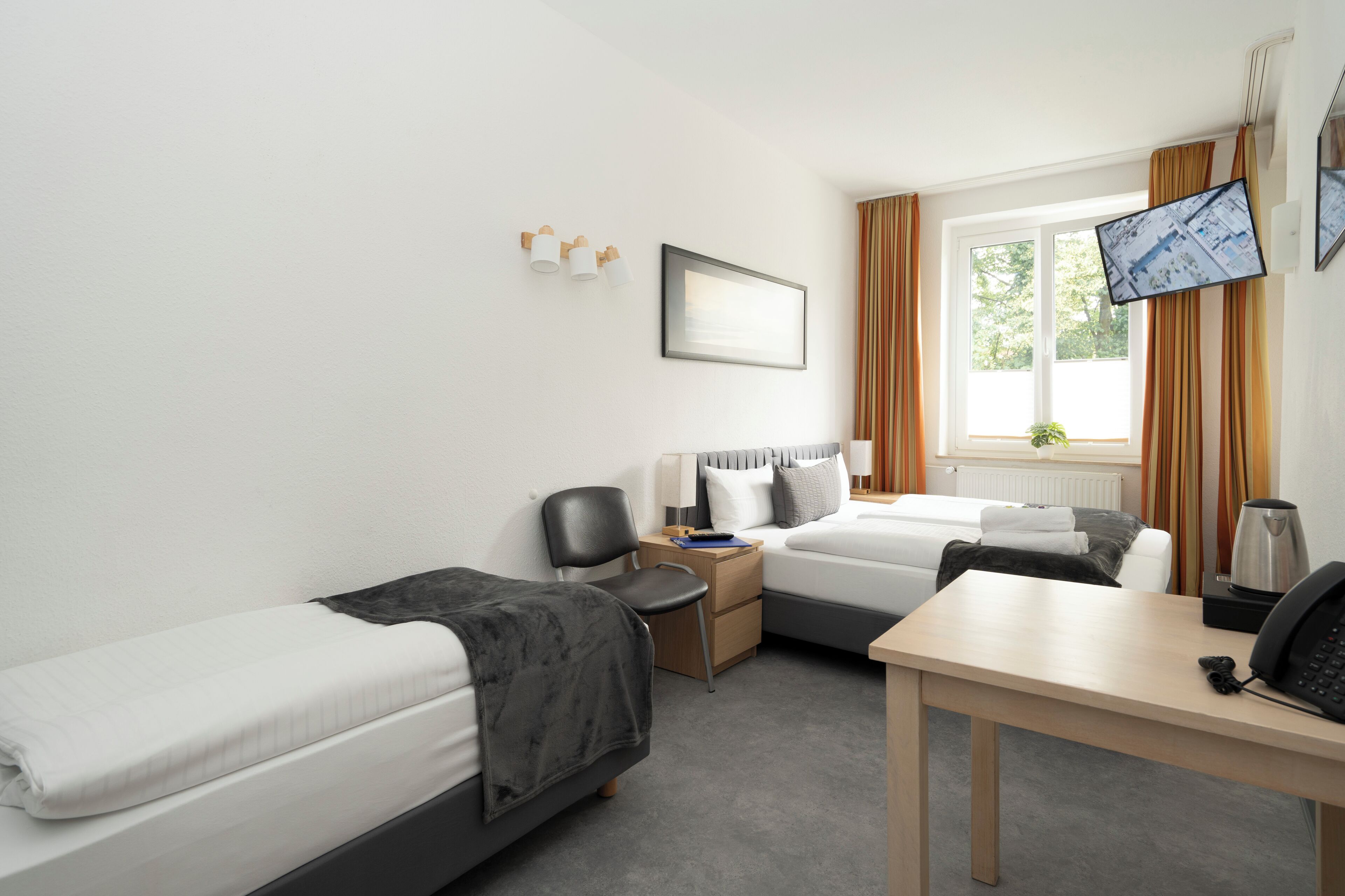 A hotel room with two single beds, a desk, a chair, a small table, a TV, and a window with curtains.