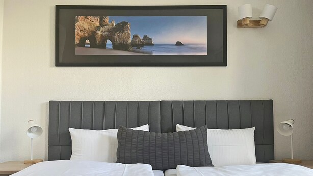 A neatly made bed with white linens, a dark headboard, and a framed coastal landscape picture above it. - Baltic Hotel (Lübeck)