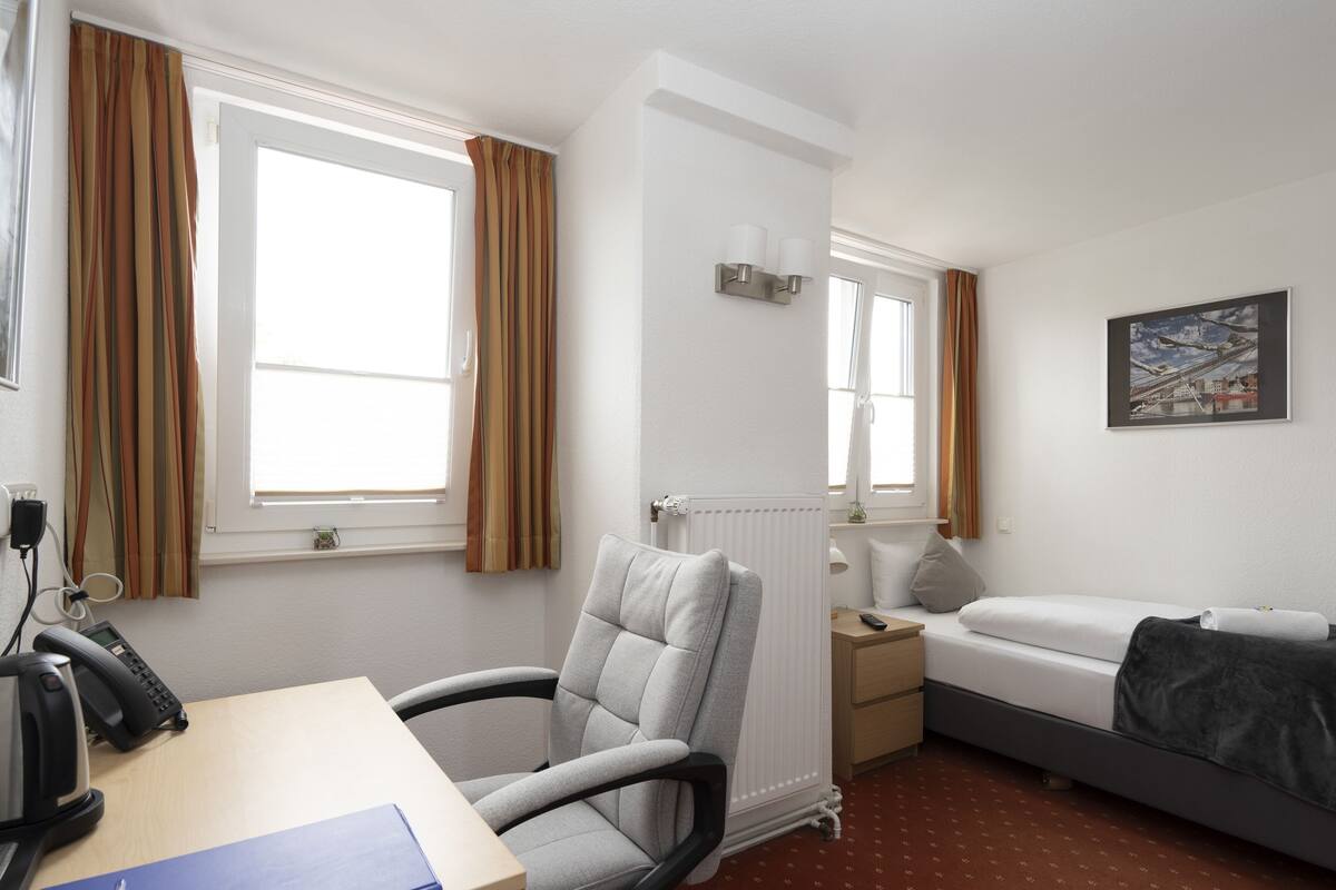 A hotel room with a bed, a desk with a telephone, a chair, a radiator, and a painting on the wall.