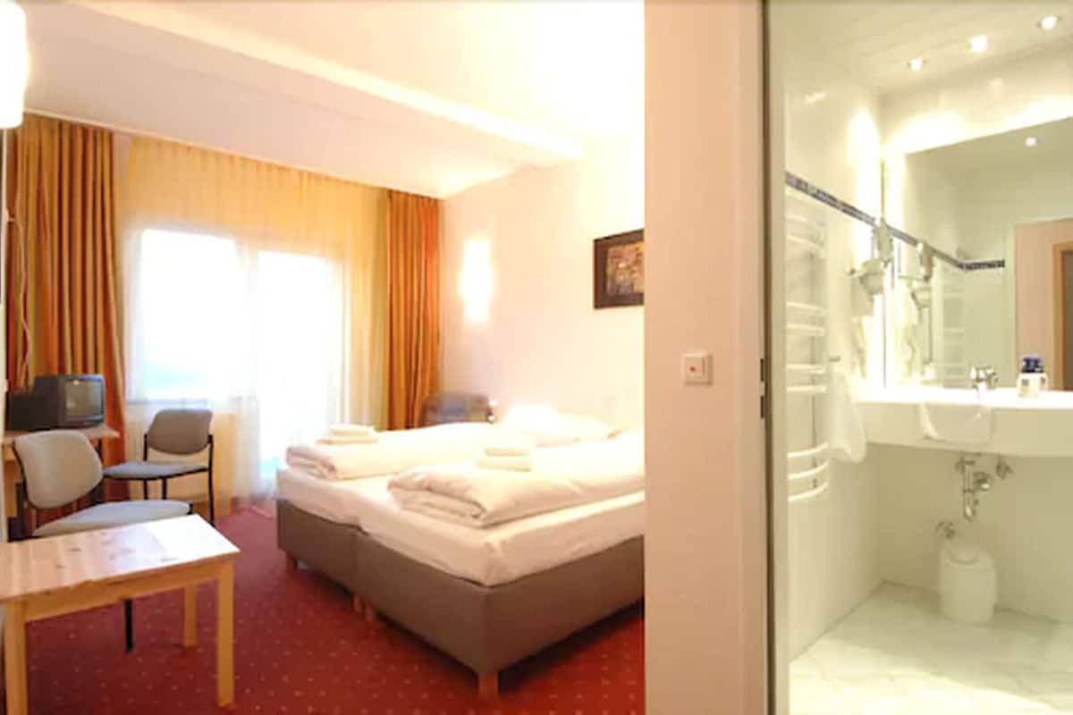 A hotel room with a bed, a desk, a chair, a television, and a bathroom with a sink and mirror.