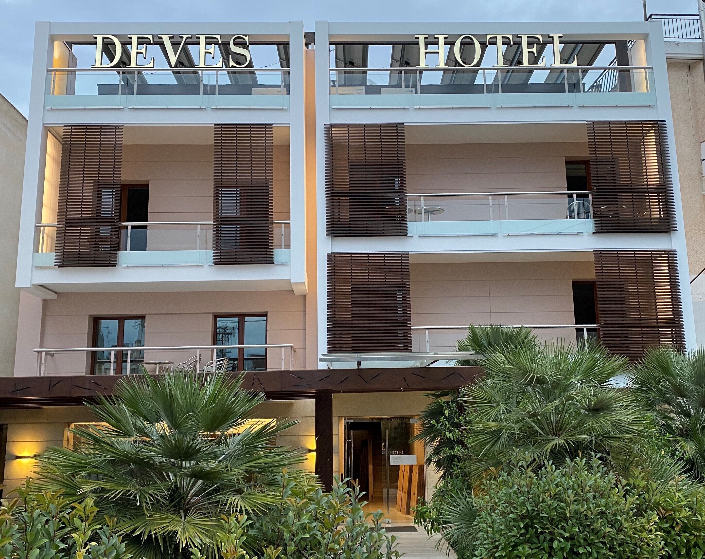 A hotel with a modern design, featuring balconies with wooden shutters, a sign reading "DEVES HOTEL", and lush greenery in front.