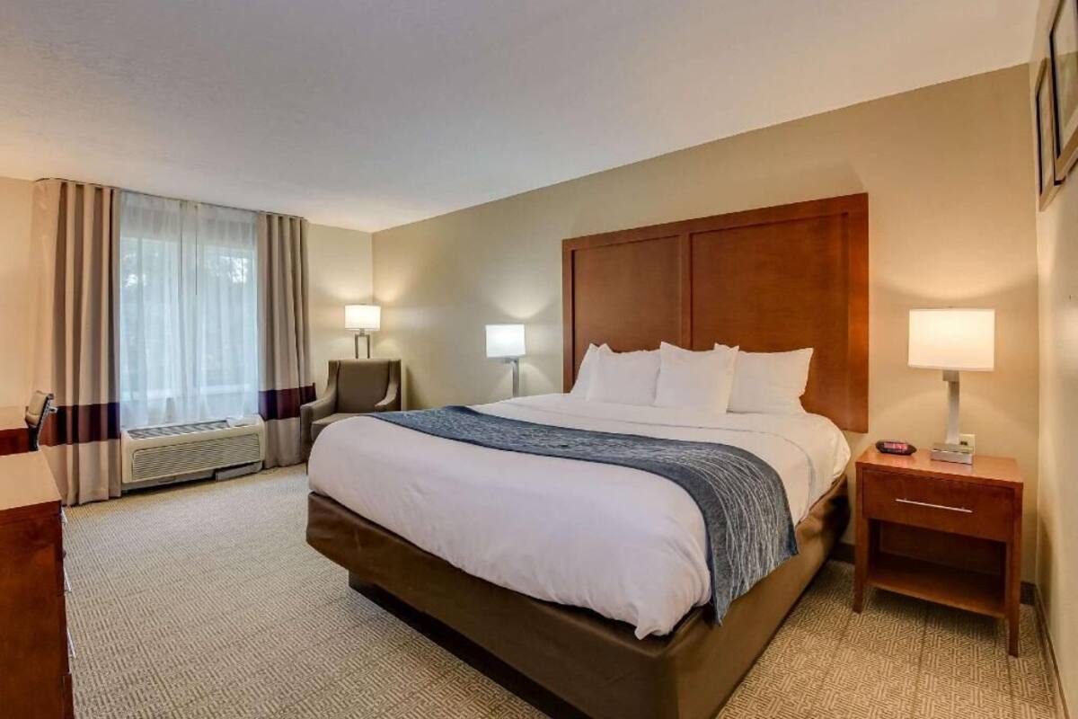 Quarto de hotel com uma cama grande, duas mesas de cabeceira com abajures, uma janela com cortinas e um aquecedor fixado na parede.