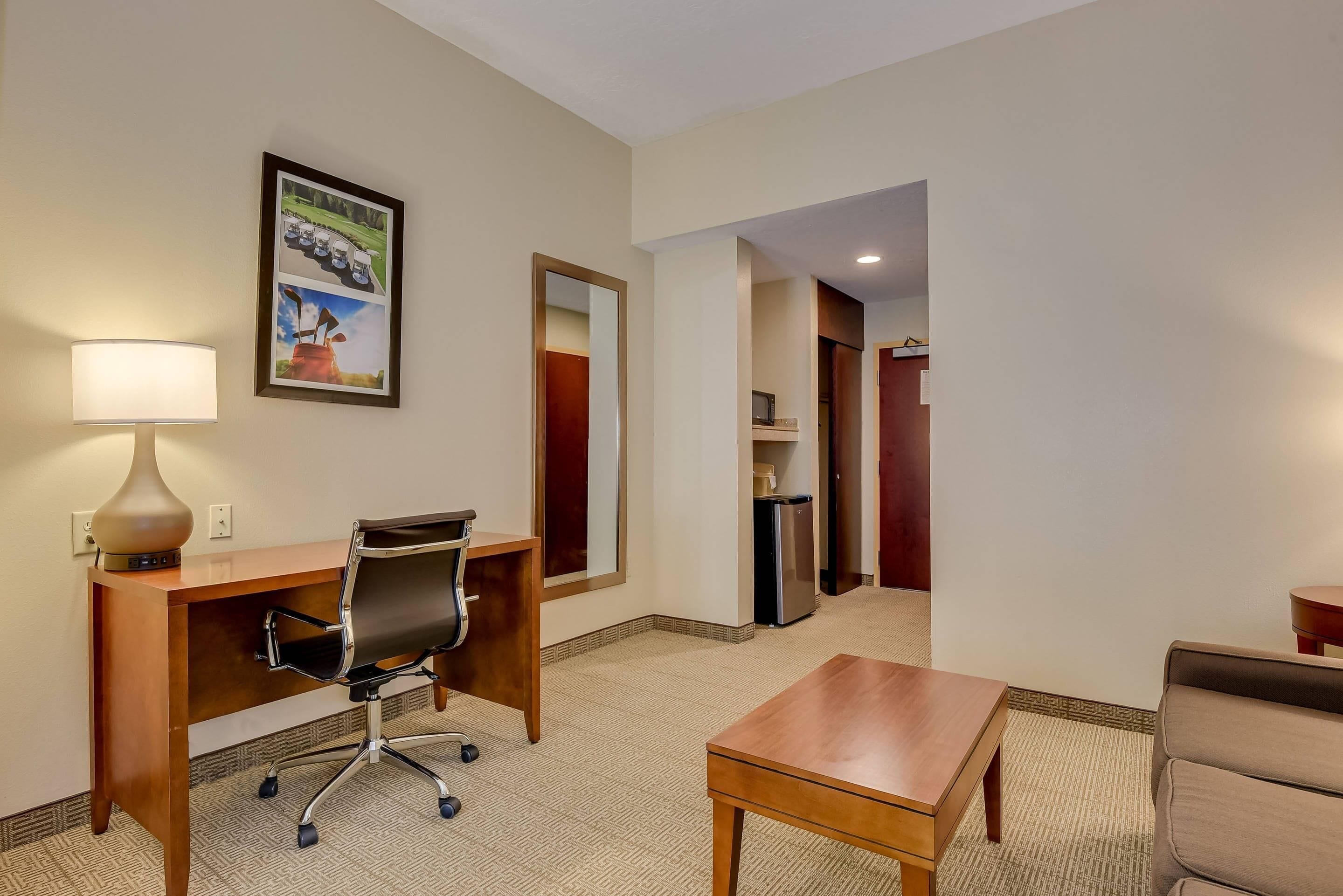 A hotel room with a wooden desk, a chair, a small table, a lamp, and a framed picture on the wall.