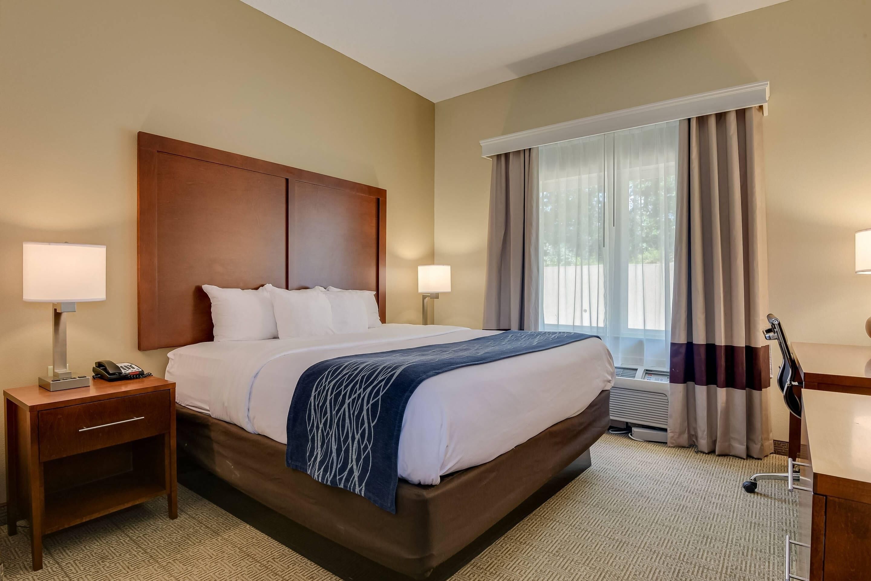 A hotel room with a large bed, two bedside lamps, a nightstand, a desk with a lamp, and a window with curtains.