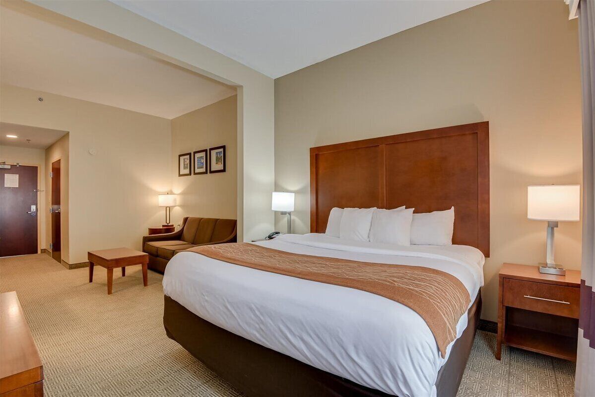 A hotel room with a large bed, a wooden headboard, a nightstand, a lamp, a small table, and a sofa.