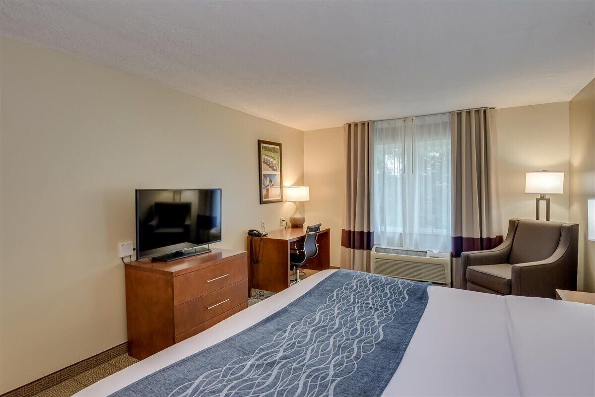 Habitació d'hotel amb llit, televisió, escriptori, cadira i finestra amb cortines.