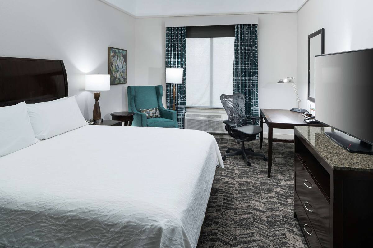 A hotel room with a large bed, a desk with a television, a chair, and a window with patterned curtains.