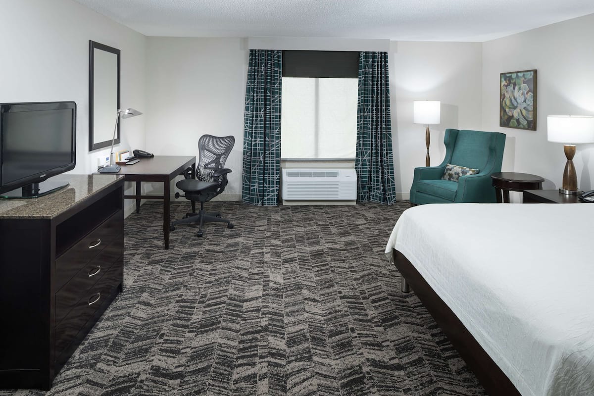 A hotel room with a bed, desk, chair, television, air conditioning unit, and a window with curtains.