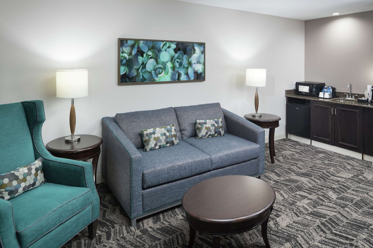 A hotel room with a grey sofa, teal armchair, and a small round table. There is a kitchen area with a microwave and coffee maker.