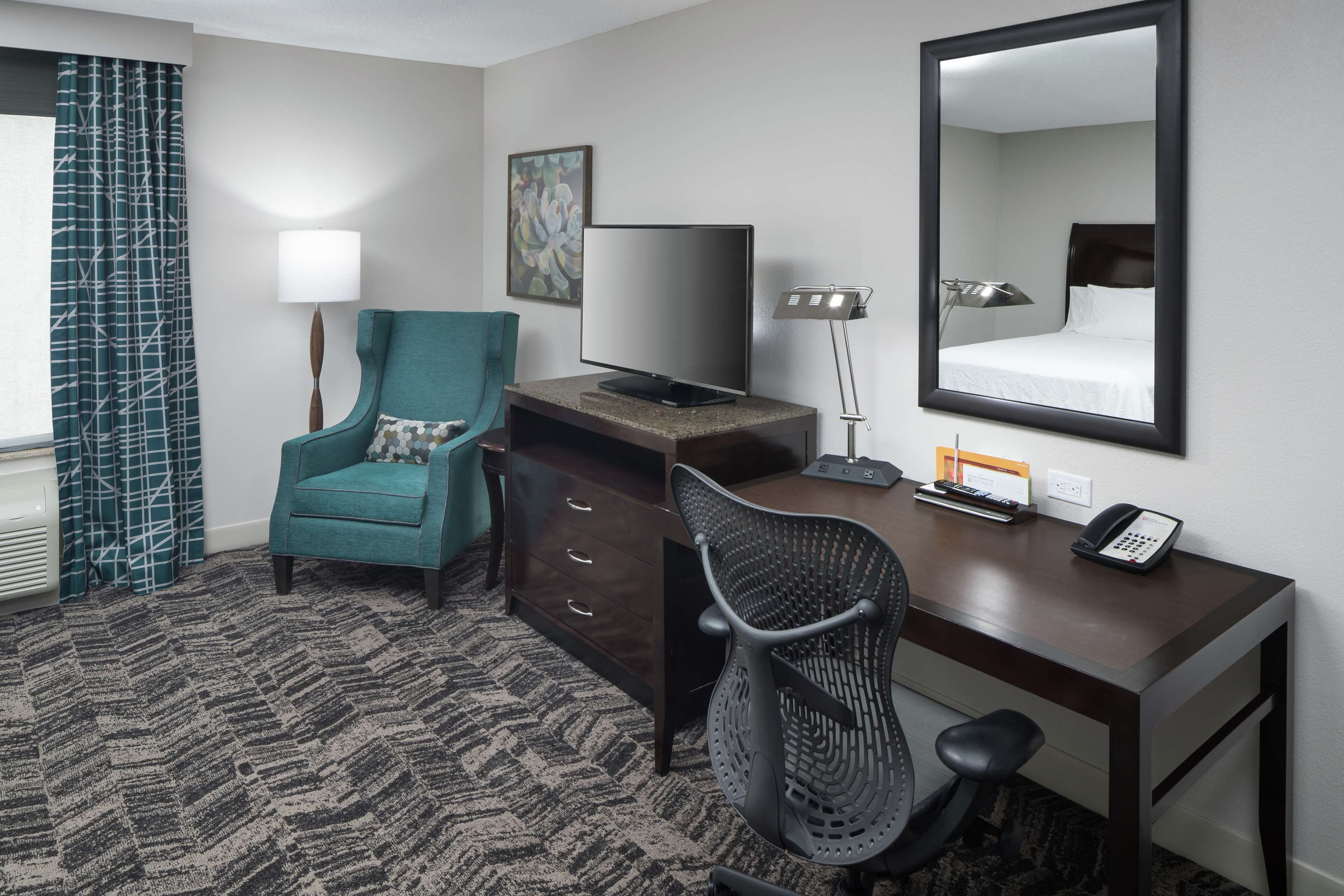 Habitación de hotel con un sillón verde azulado, un escritorio con una computadora, un televisor de pantalla plana, un teléfono y un espejo grande.