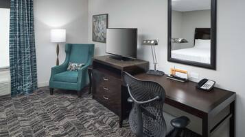 Habitación de hotel con un sillón verde azulado, un escritorio con una computadora, un televisor de pantalla plana, un teléfono y un espejo grande.