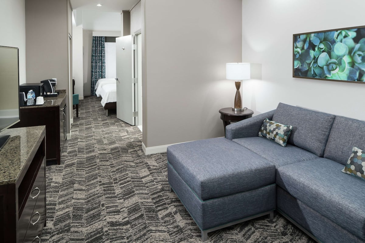 A hotel room with a gray sectional sofa, a small side table with a lamp, a large abstract painting on the wall, a dark wooden dresser, a bed with white linens, and a patterned carpet.