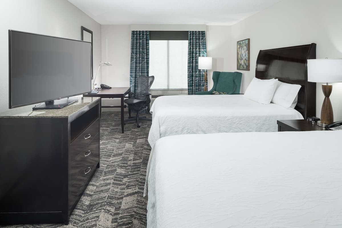 A hotel room with a large flat-screen TV, a desk with a chair, two beds, a desk lamp, and a painting on the wall.