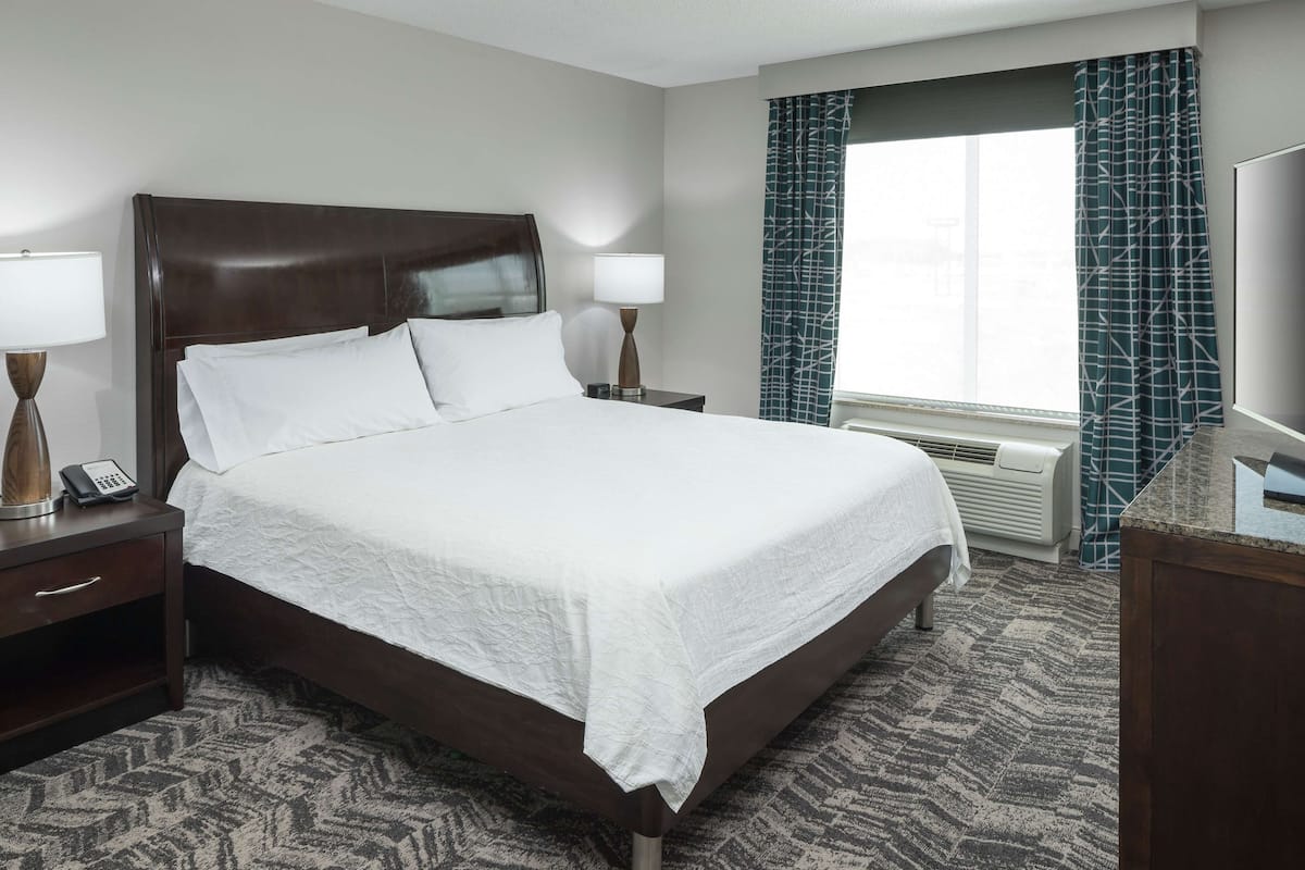 A hotel room with a large bed, two bedside tables with lamps, a telephone, and a window with curtains.