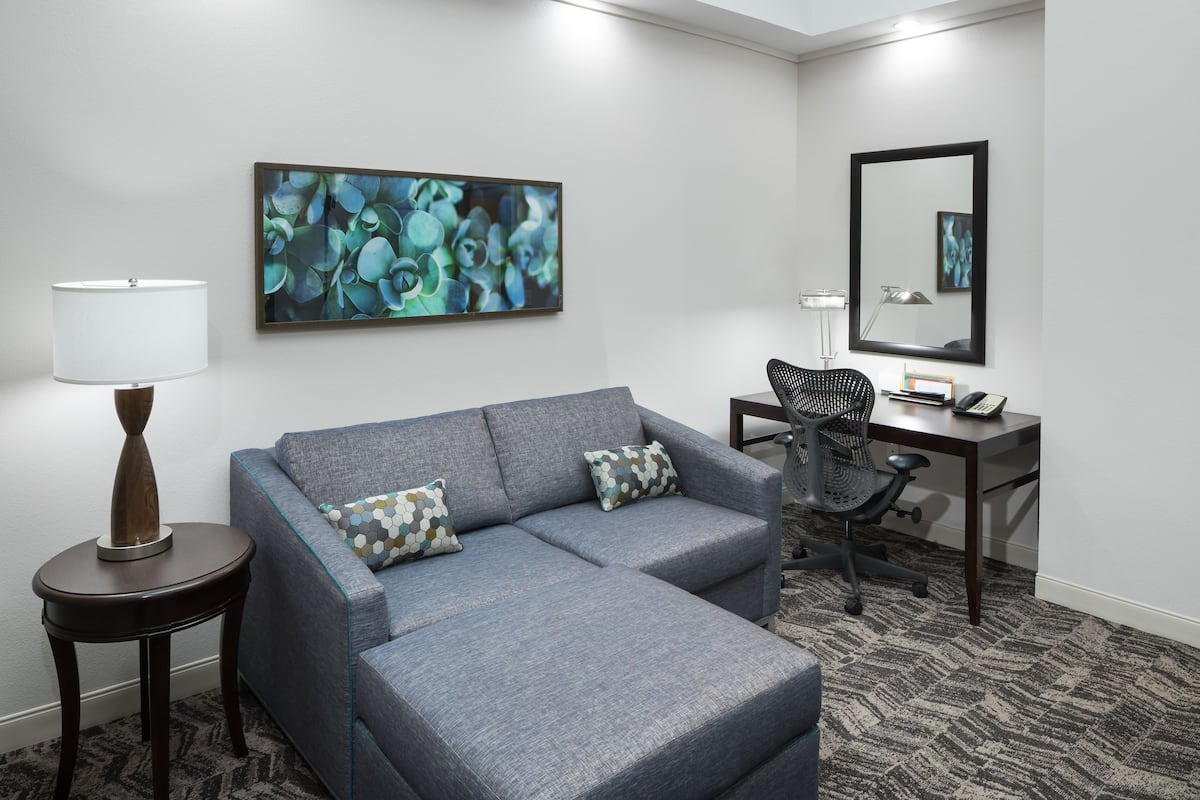A hotel room with a grey sofa, a small side table with a lamp, a desk with a chair, a large abstract painting, and a mirror on the wall.