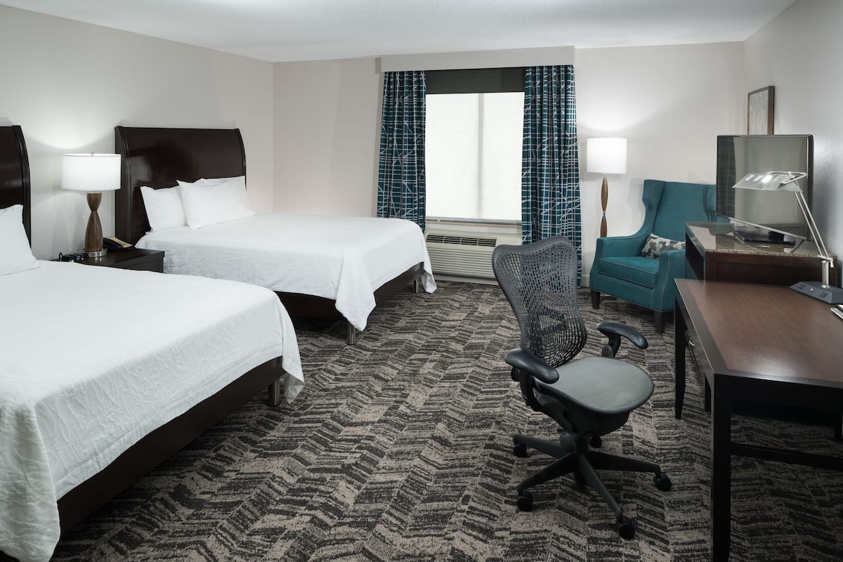 A hotel room with two beds, a desk, a chair, a TV, and a window with curtains.