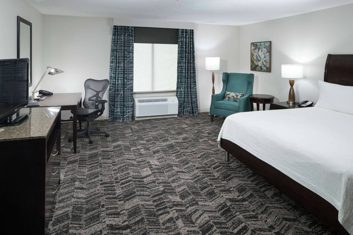 A hotel room with a bed, desk, chair, TV, air conditioning unit, and a window with curtains.