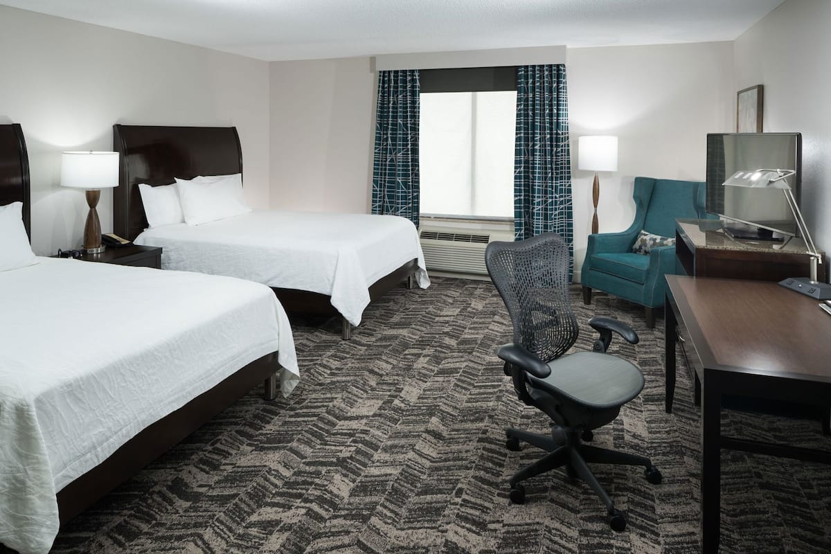 A hotel room with two beds, a desk, a chair, a lamp, a window with curtains, and a wall-mounted TV.