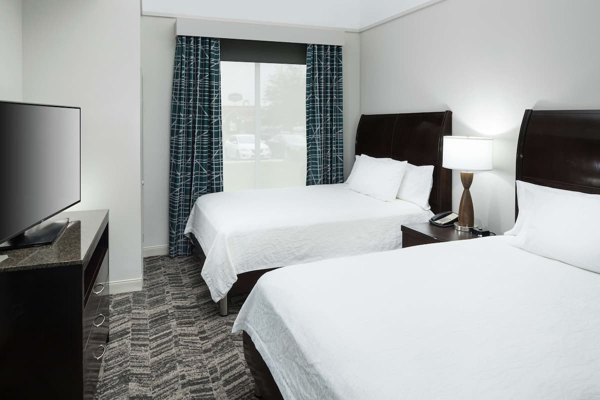 A hotel room with two beds, a TV, a desk, and a window with curtains.