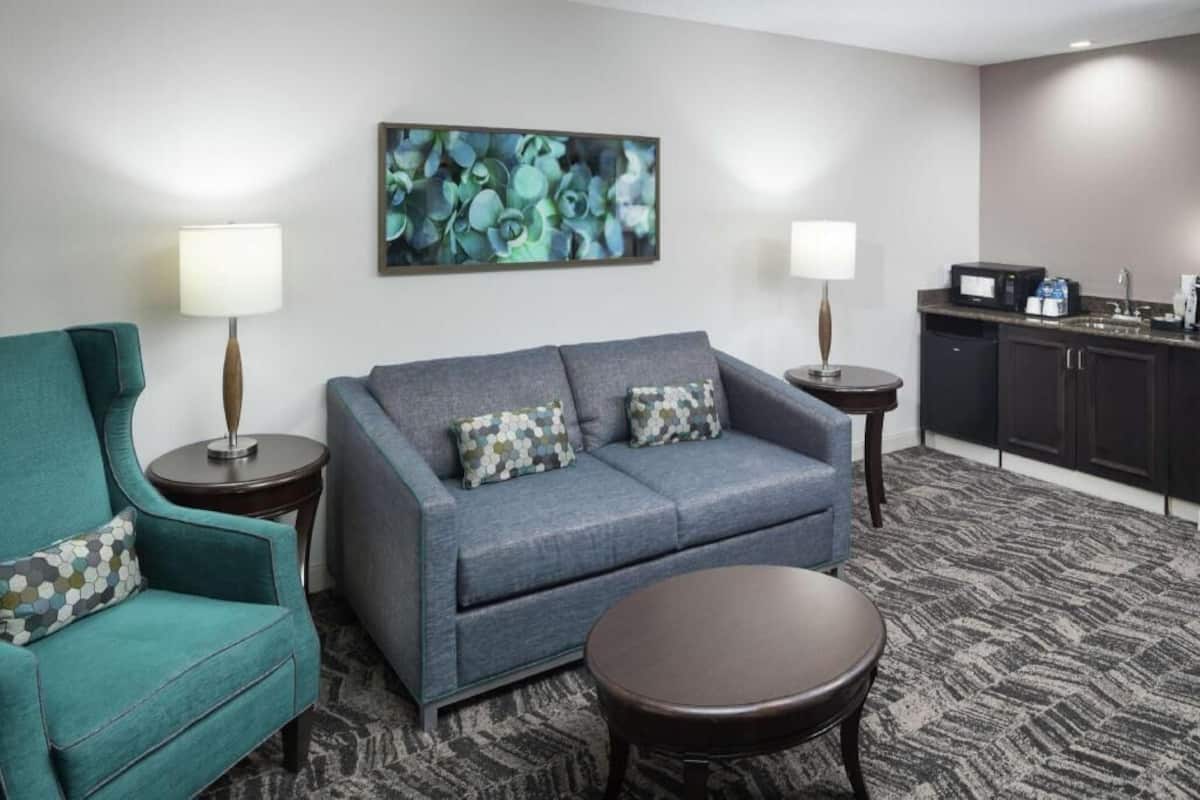 A hotel room with a grey sofa, teal armchair, and a small round table.