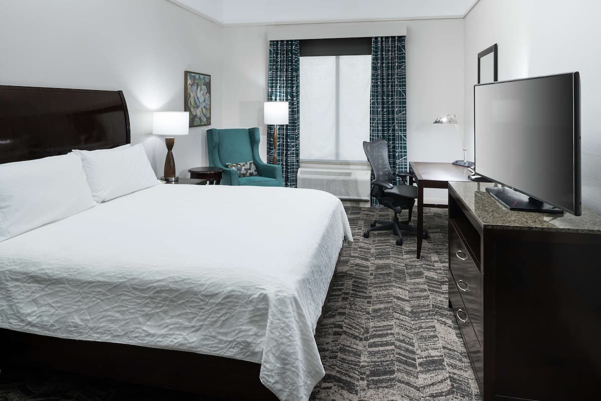 A hotel room with a large bed, a desk with a computer, a TV, and a view of the outdoors through a window with curtains.