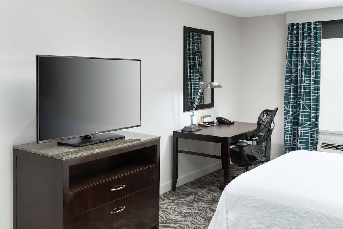 A hotel room with a bed, a desk with a chair, a television on a cabinet, a desk lamp, and a mirror.