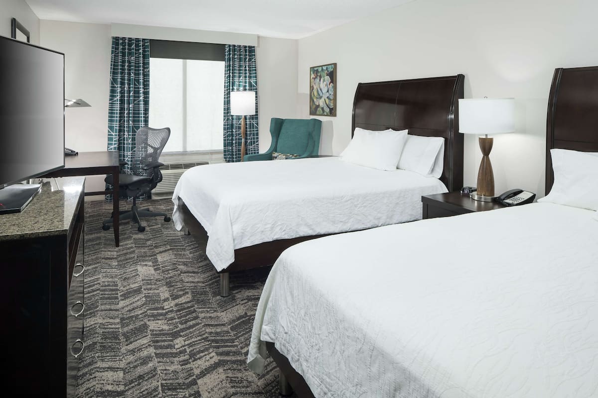 A hotel room with two beds, a desk, a TV, and a chair.